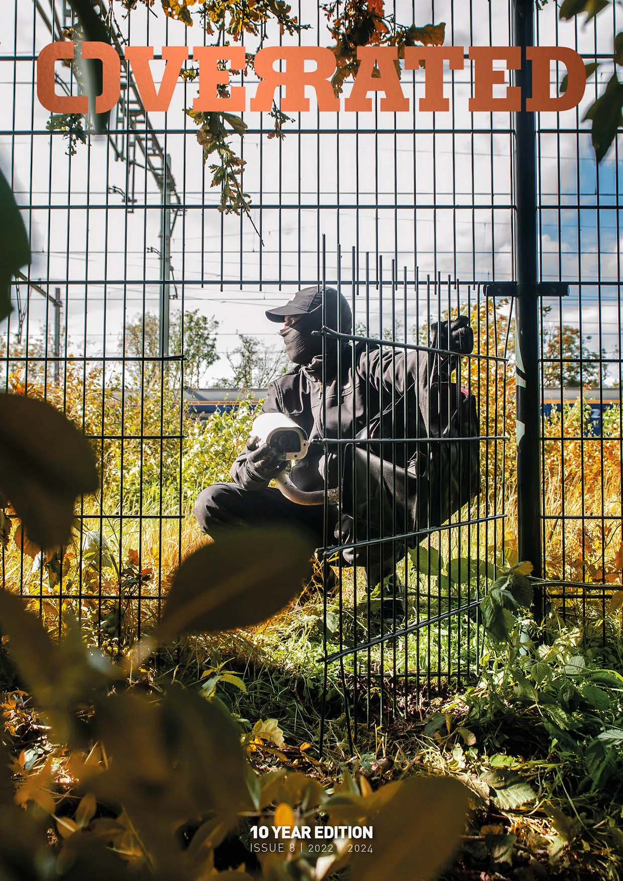 The cover of Overrated Magazine issue 8, showing a person sitting behind a cut-open fence, holding a security camera 